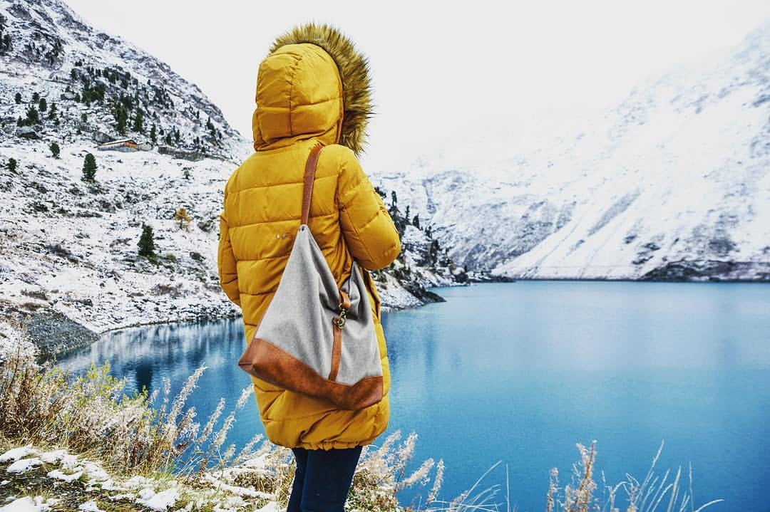 woman in a yellow parka with a handbag in front of a lake and snow covered mountains